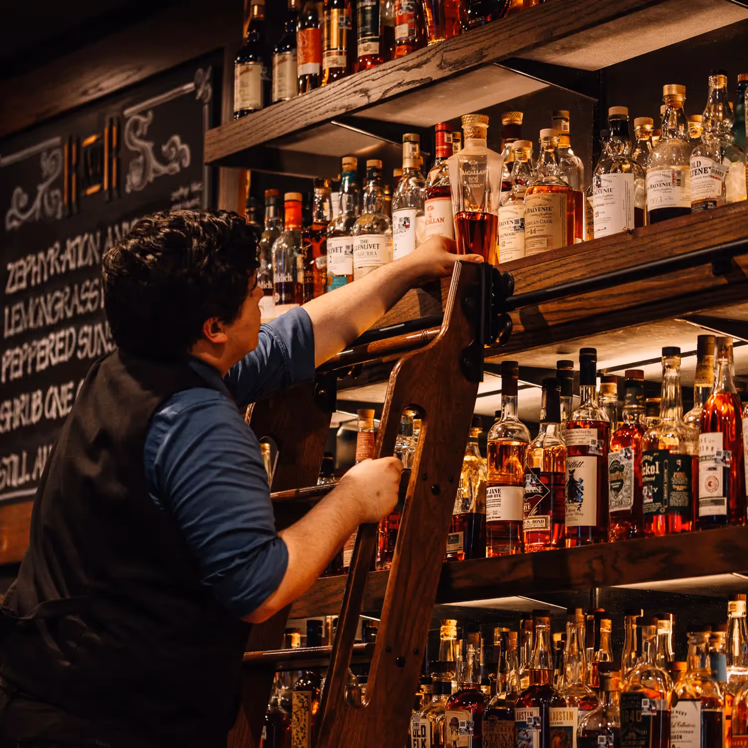 A Rock and Rye bartender on the rolling library ladder, reaching for a bottle on the top shelf